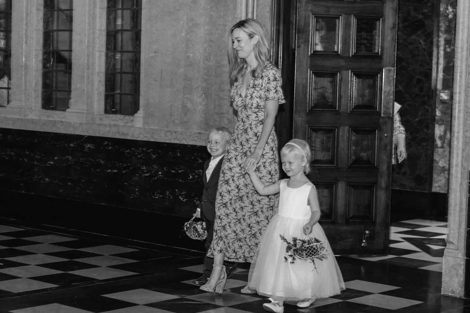 A woman and two children walking inside a building. The woman is holding the hand of the young girl and carrying a small bag, both children are smiling. The girl wears a white dress and carries a basket of flowers; the boy is dressed in a suit.