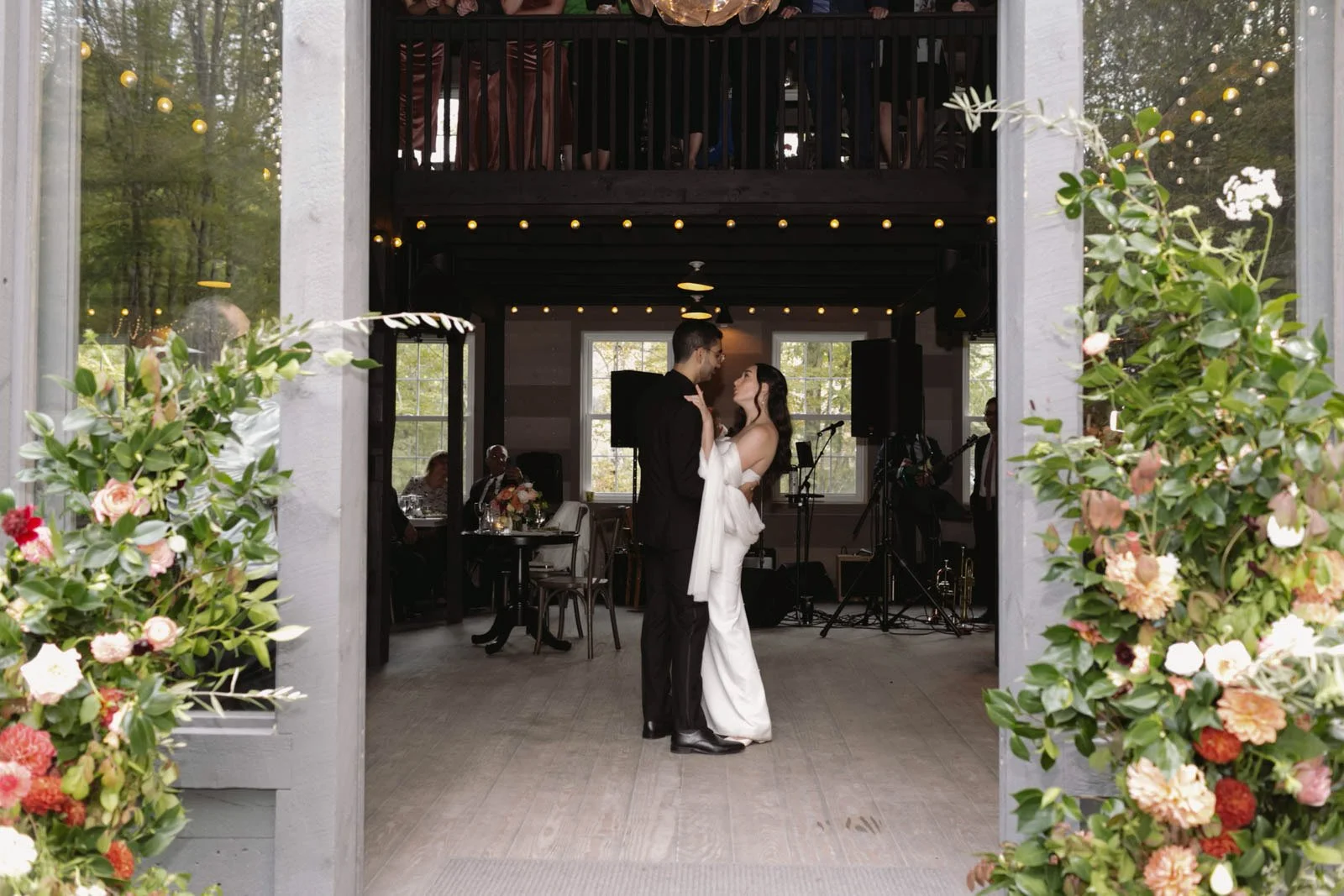 A couple dancing at a wedding reception inside a decorated venue, with guests seated at tables, band equipment, and floral arrangements surrounding the entrance.