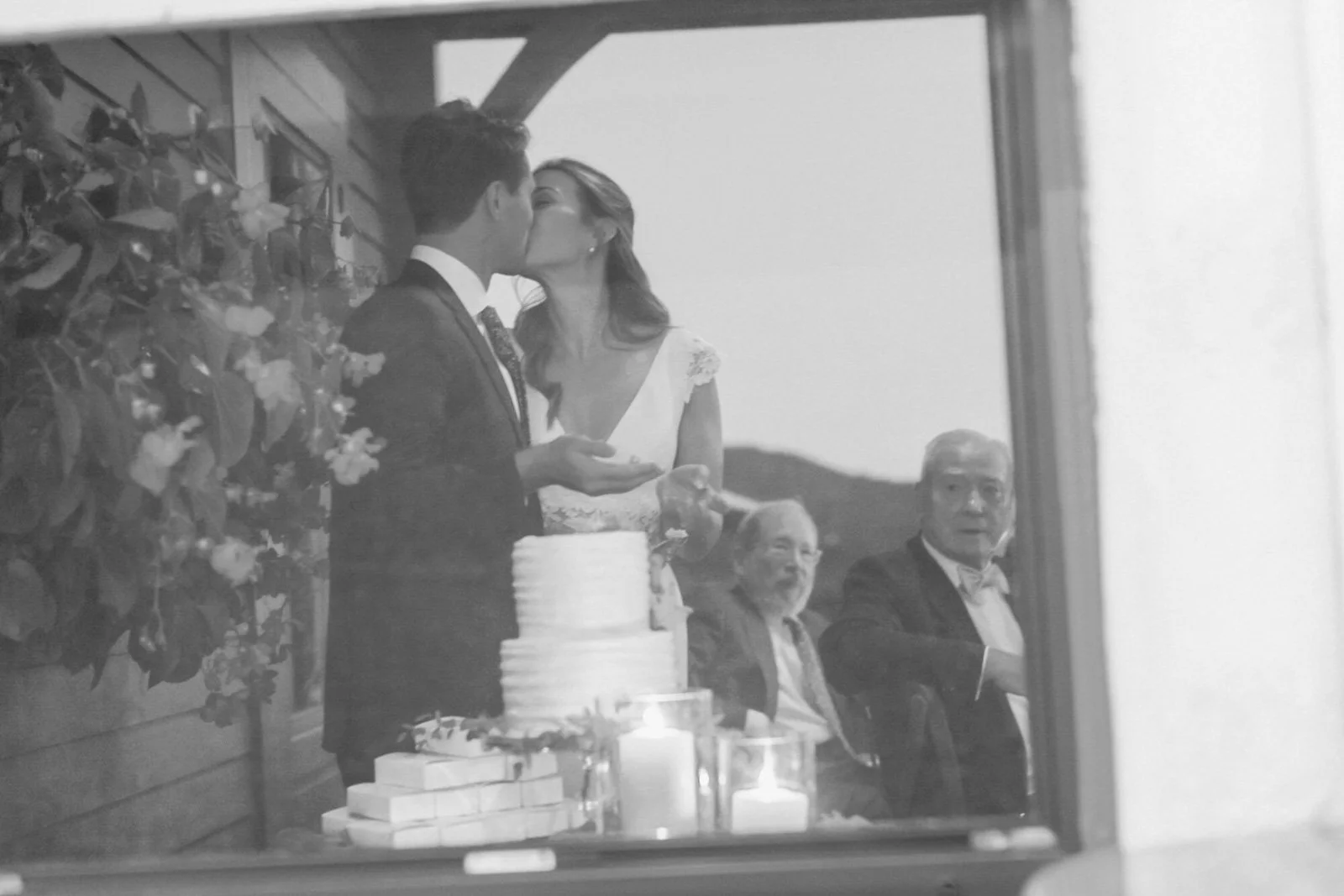 Black and white photo of a wedding reception showing a couple kissing, a wedding cake, and three older men seated at the table.