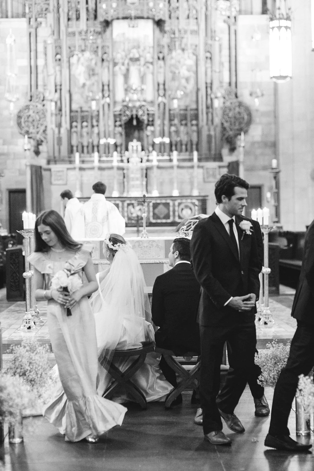 A black-and-white photo of a wedding ceremony inside a church with a high altar, candles, and religious decorations. The bride, wearing a wedding veil, and the groom, in a suit, are seated at the altar. Several guests and a flower girl holding flower