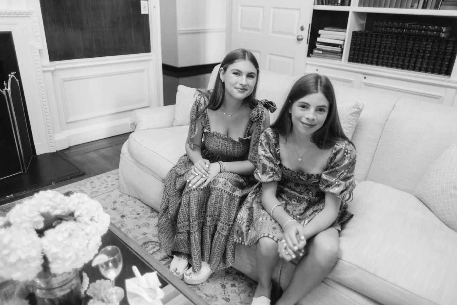 Two young girls sitting on a sofa in a living room, smiling at the camera. The room has bookshelves and a door in the background, and a table with flowers and a glass of drink in the foreground.