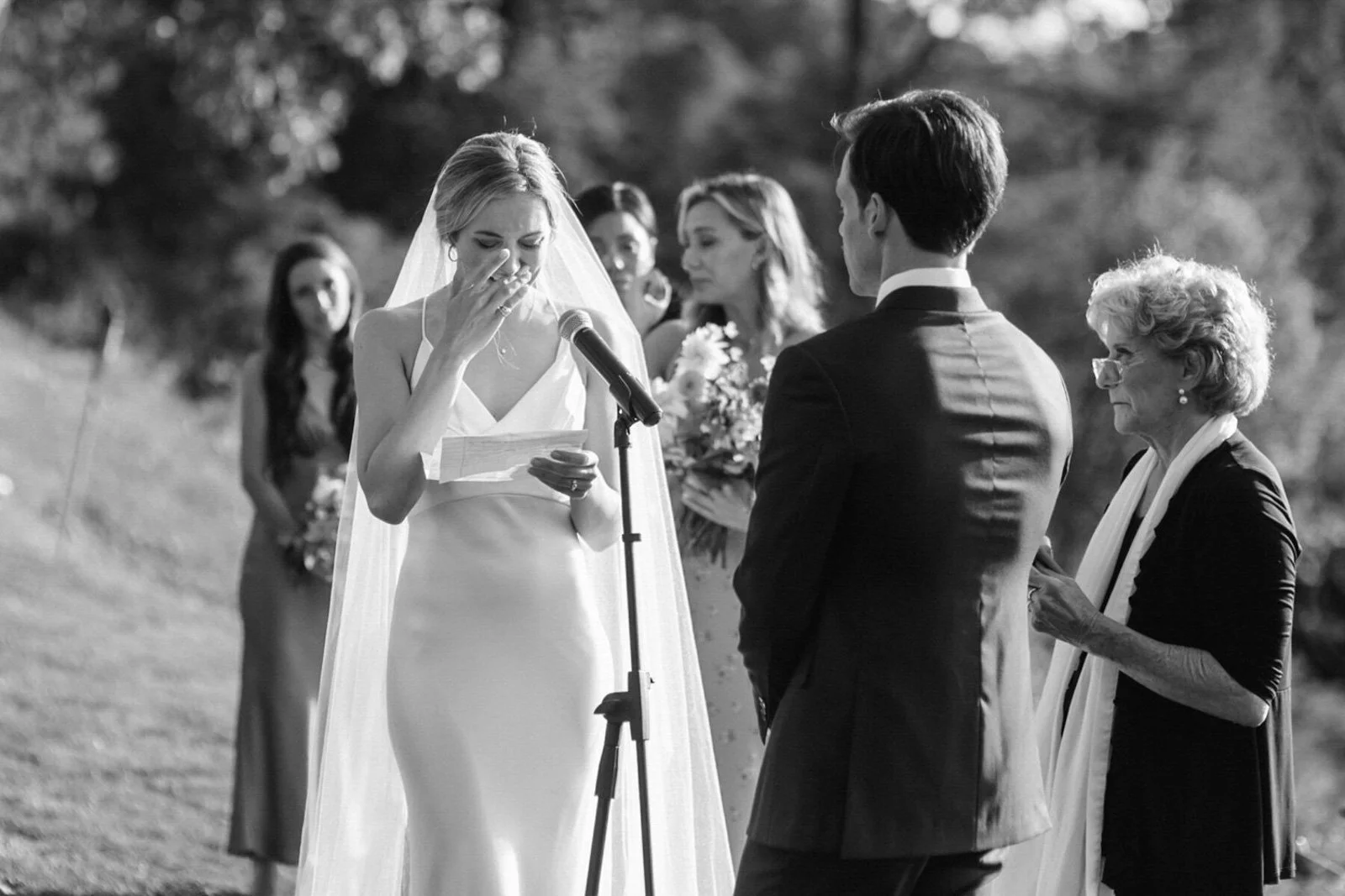 A black and white photo of a wedding ceremony outdoors with a bride reading a vow or speech at a microphone, a groom facing her, and three women witnessing, including an older woman with glasses and a white scarf.
