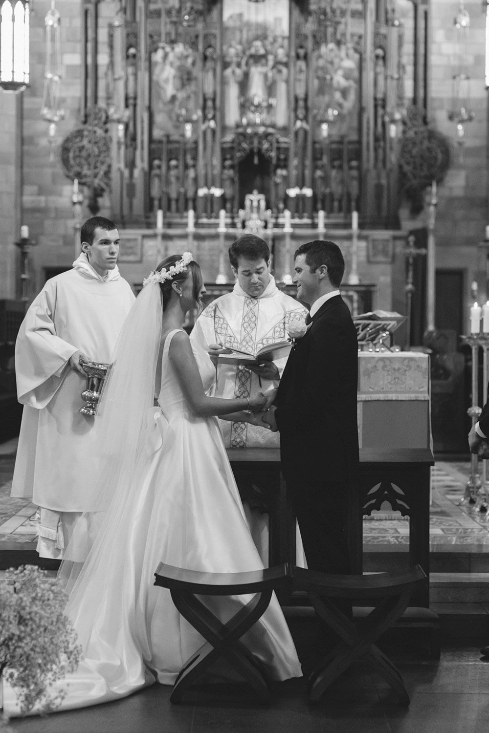 A black-and-white photo of a wedding ceremony inside a church. The bride and groom are holding hands, facing each other, with a priest officiating the ceremony. An altar with candles and religious artwork can be seen in the background.