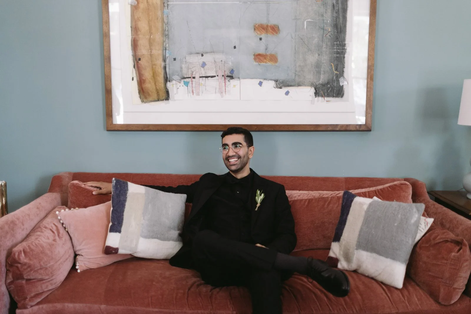 A man with glasses and a boutonniere sits on a reddish-brown sofa, smiling and relaxed, with a light blue wall, a large abstract painting, and a white table lamp in the background.