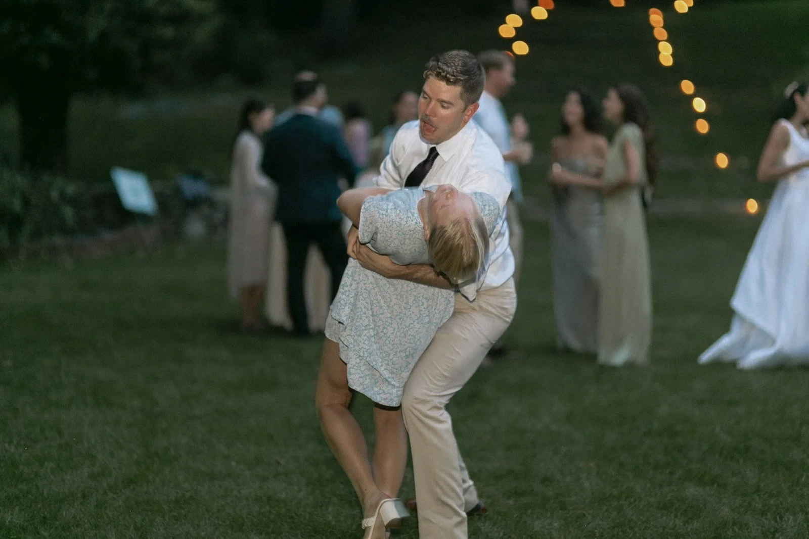 A man in a white shirt and beige pants is dancing with an older woman in a gray dress at an outdoor evening event. The man is holding the woman in a dip, and there are other people in the background socializing and enjoying the event.