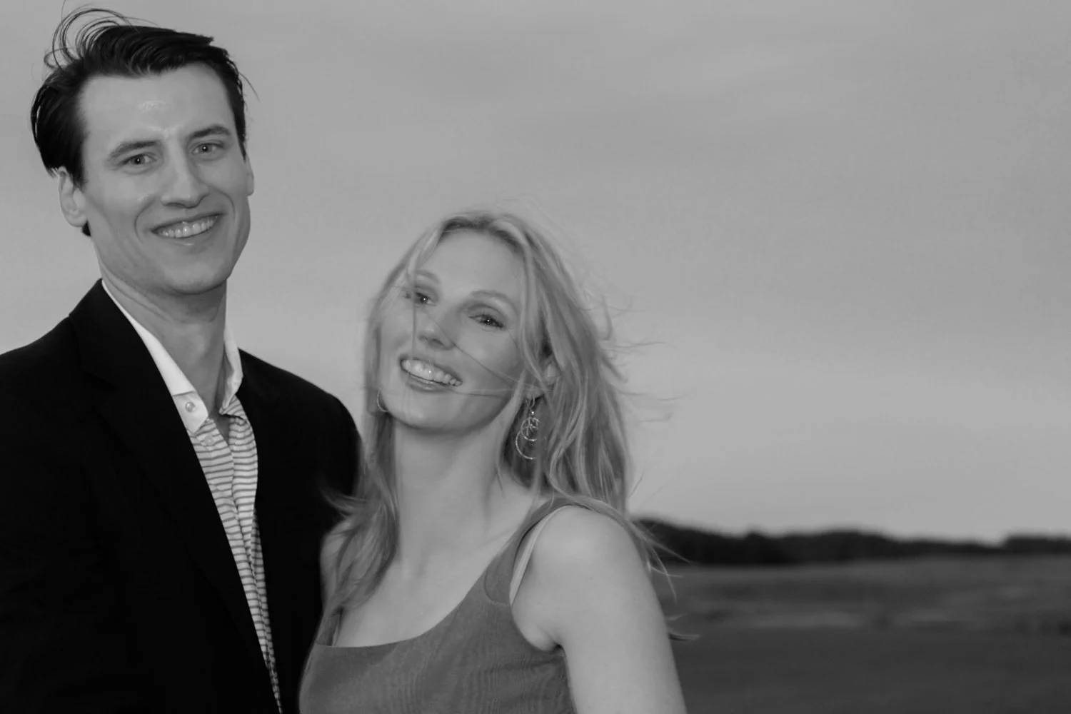 A smiling man and woman standing outdoors, with an open field and cloudy sky in the background.