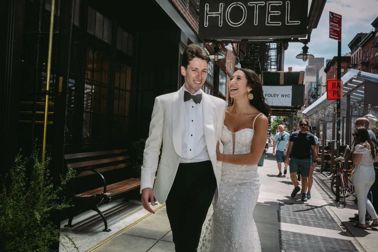 A smiling newlywed couple in wedding attire walking on a city sidewalk, surrounded by pedestrians under city awnings and signs.