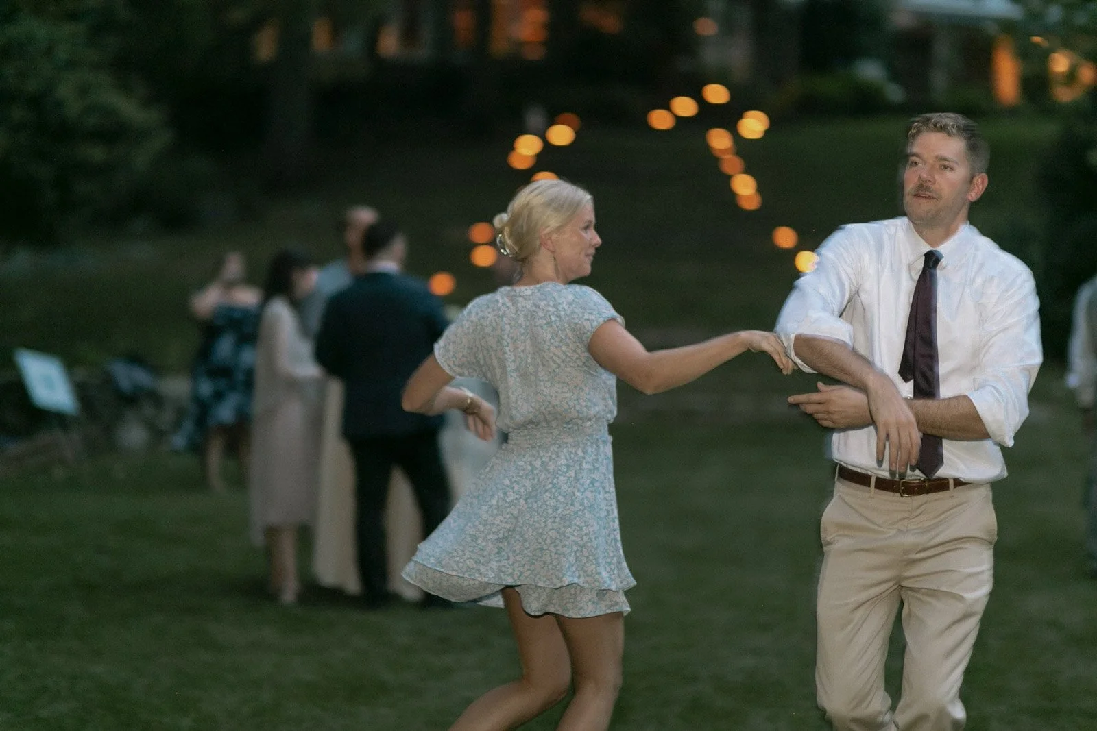A woman in a blue floral dress dancing with a man in a white shirt and tan pants at an outdoor evening event. Other guests are in the background, and string lights are visible.
