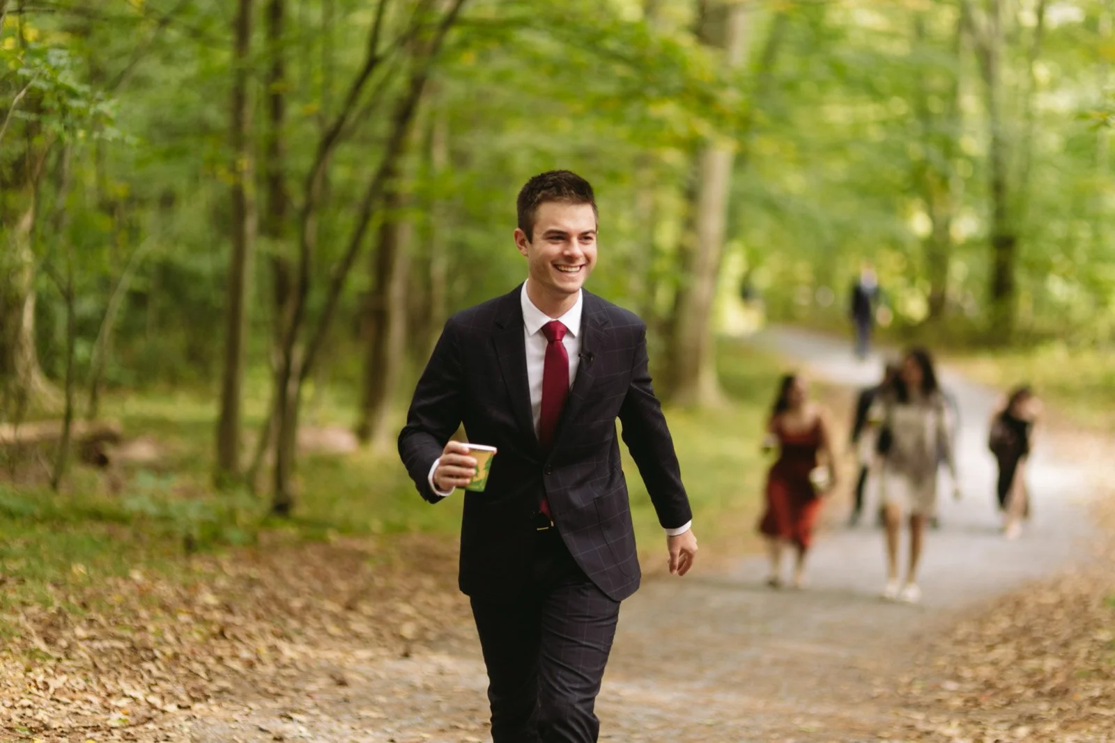 A young man in a formal suit with a red tie smiling and walking in a wooded park, holding a coffee cup. Several other people are walking in the background.