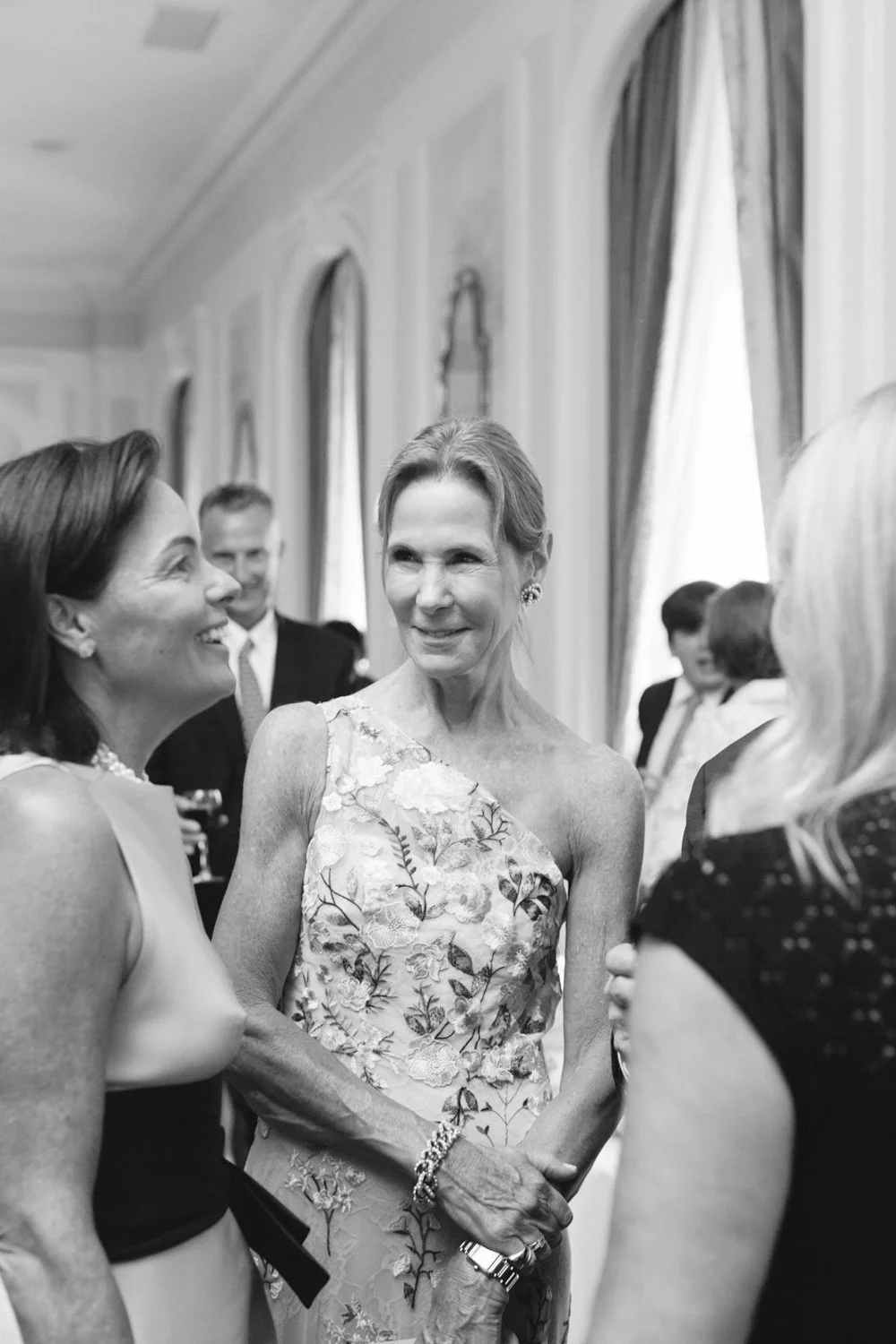 Black and white photo of three women at a social gathering, smiling and engaging in conversation indoors with elegant curtains and wall decor in the background.