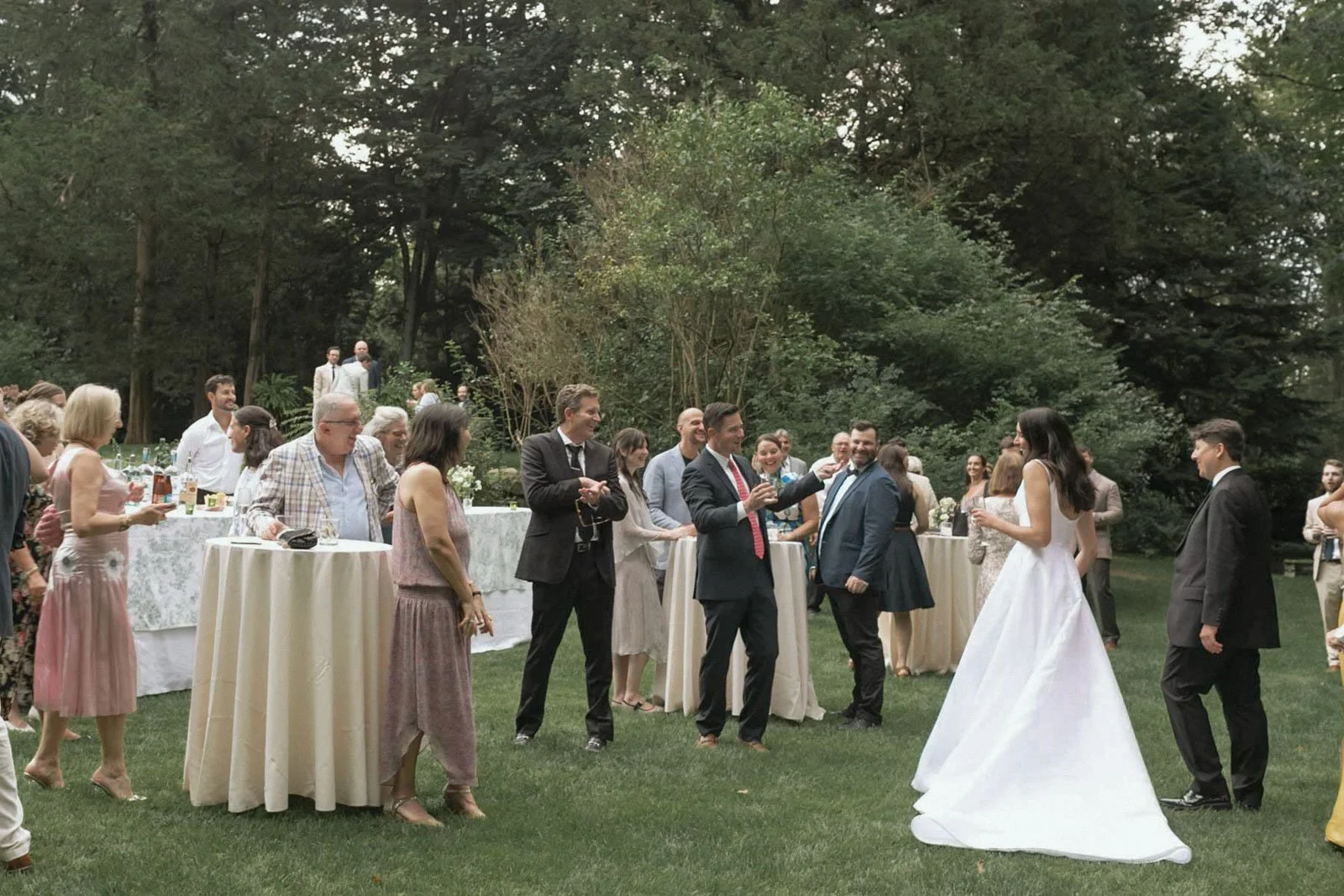 People gathered outdoors at a wedding reception near tables with drinks, trees, and guests socializing, with the bride and groom in the foreground.