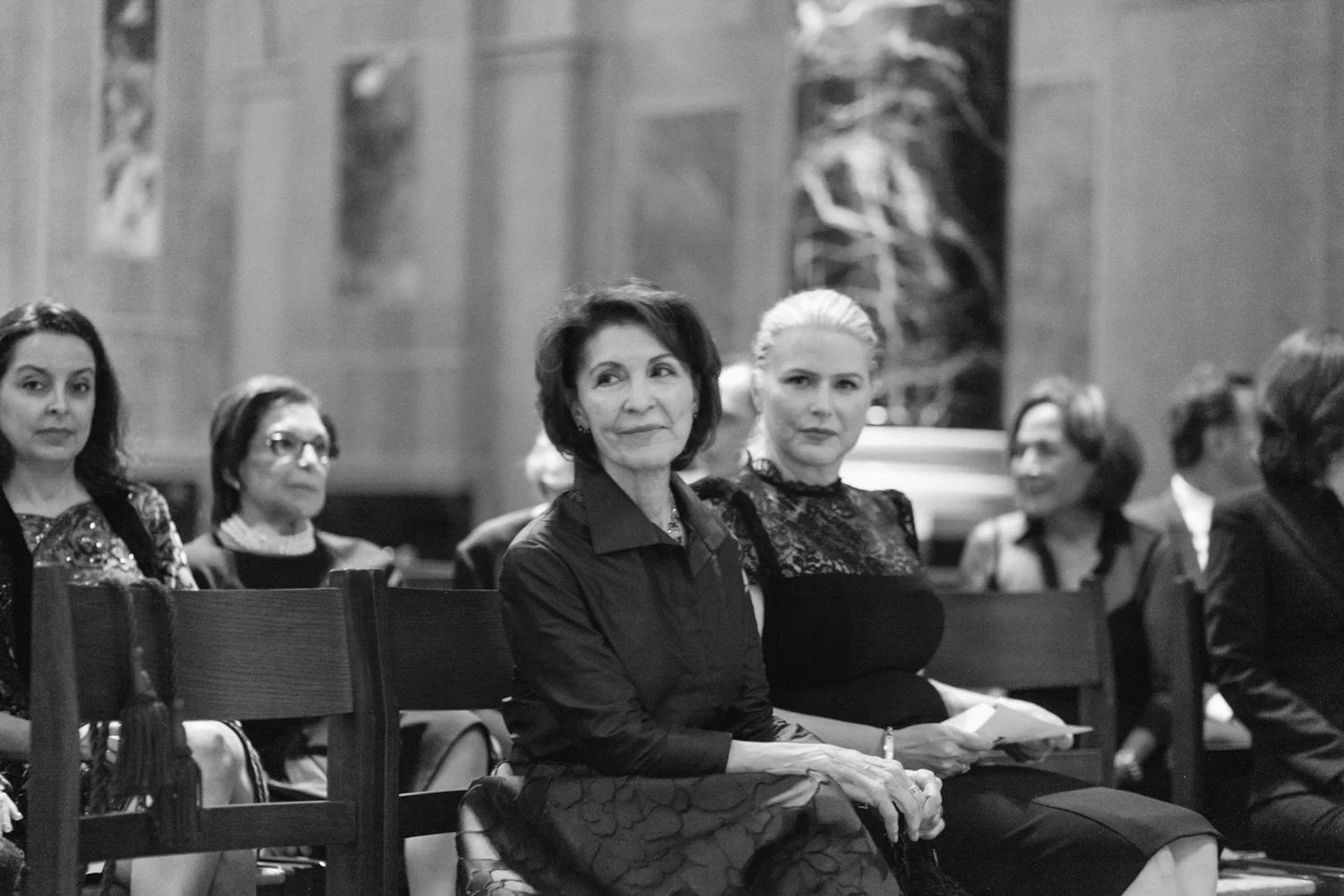 Black and white photo of women seated in a row indoors, wearing formal attire, at what appears to be a formal event or gathering.