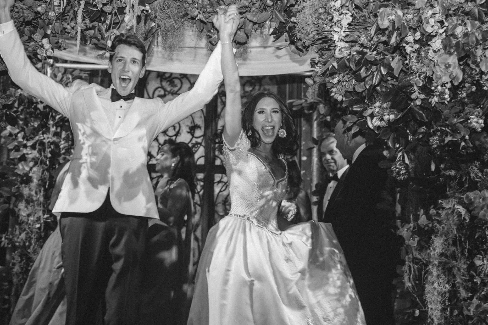 A joyful wedding couple celebrating under a floral arch, with the groom raising his arm and the bride holding his hand, surrounded by guests.