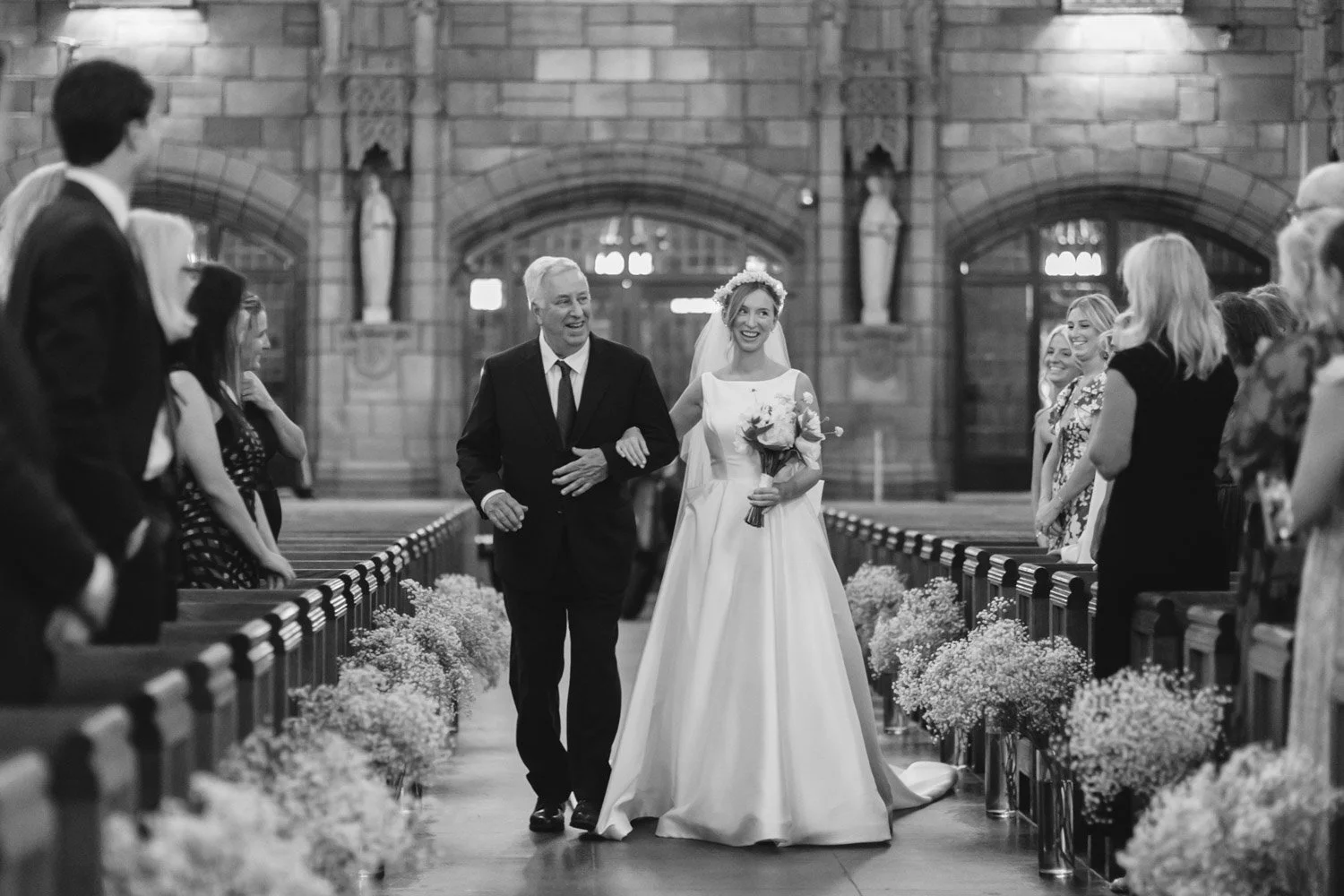 Black and white photo of a bride walking down the aisle of a church, smiling, holding a bouquet, with an older man (possibly her father) escorting her, surrounded by wedding guests.