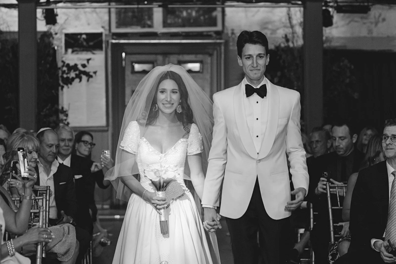 Black and white photo of a bride and groom walking down the aisle together at a wedding ceremony, holding hands, with guests seated on either side taking photos.