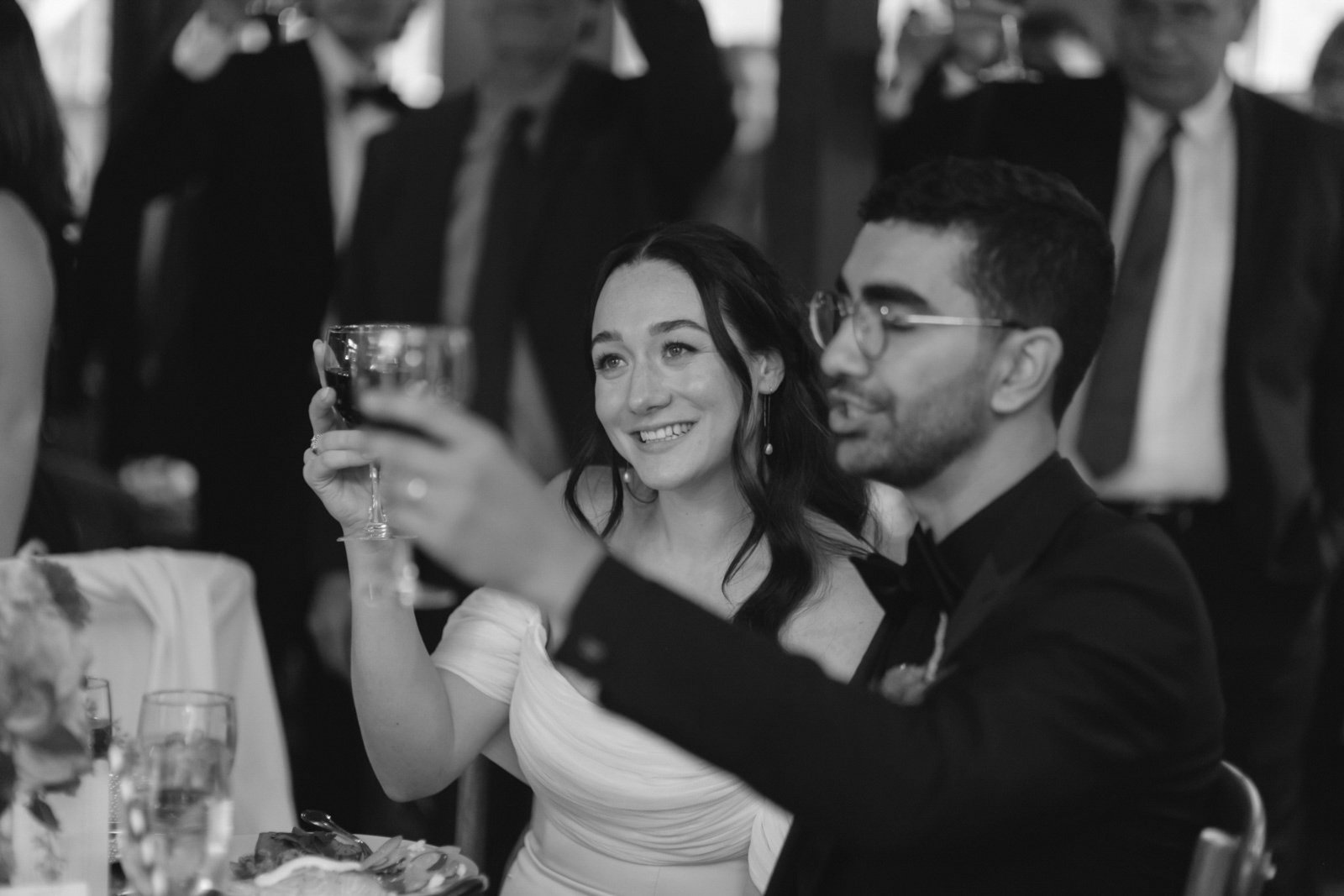A woman in a wedding dress and a man in a tuxedo with glasses are celebrating at a wedding reception, raising glasses for a toast.