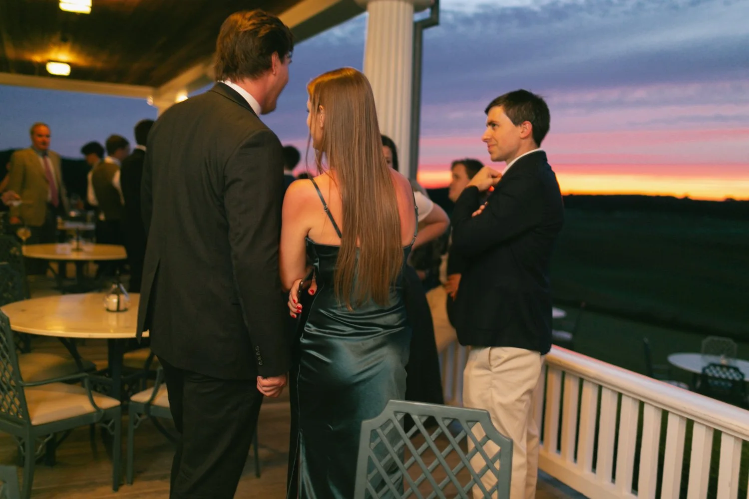 People socializing at a rooftop party during sunset, some standing near a railing, with a sunset and clouds in the background.