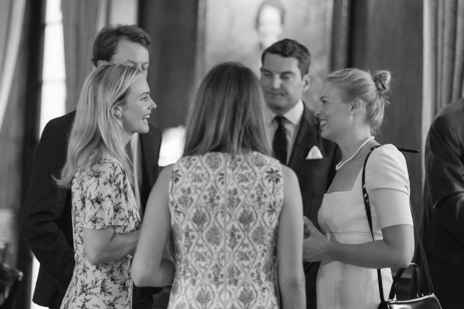 Group of five people in formal attire talking and smiling in a room, possibly at a social or professional gathering.