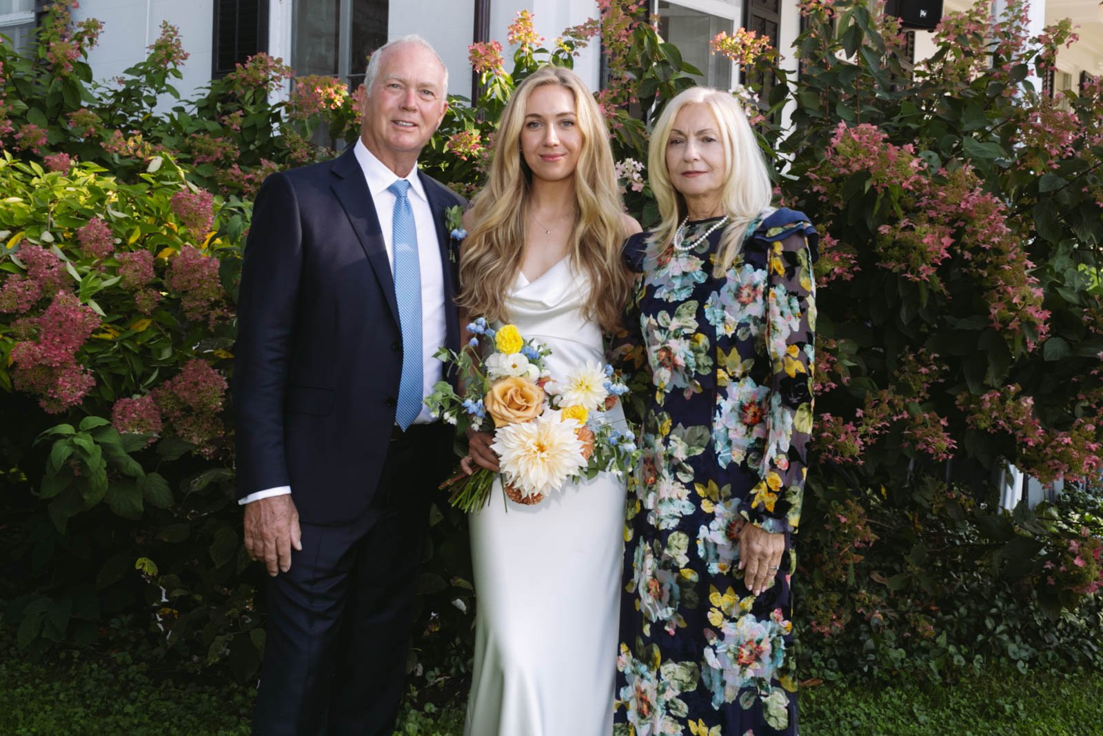 Three people standing outdoors in front of pink flowering bushes, two women and one man, dressed formally, with the woman in a white dress holding a bouquet of flowers.