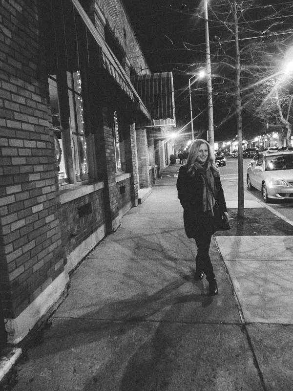 A woman with long hair walking on a city sidewalk at night, illuminated by streetlights, with parked cars and buildings in the background, in black and white.