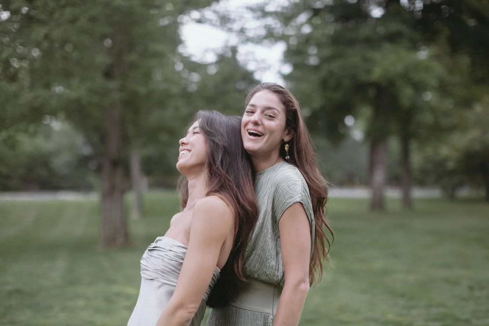 Two women standing outdoors in a park with trees in the background, smiling and enjoying each other's company.