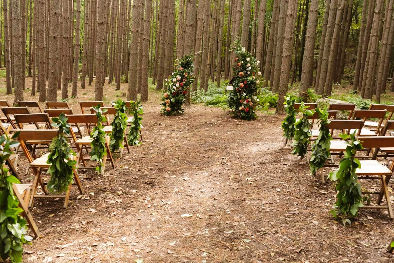 Outdoor wedding ceremony setup in a wooded area with chairs decorated with green leaves, a floral arch, and a flower arrangement at the front.