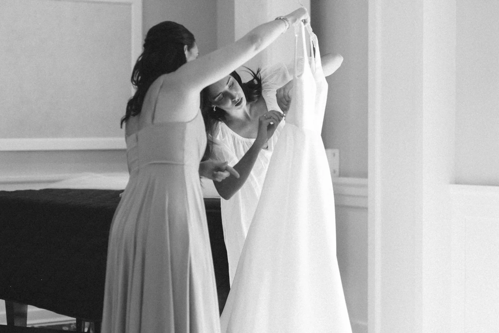 Two women in dresses examining a wedding gown on a mannequin in a room.