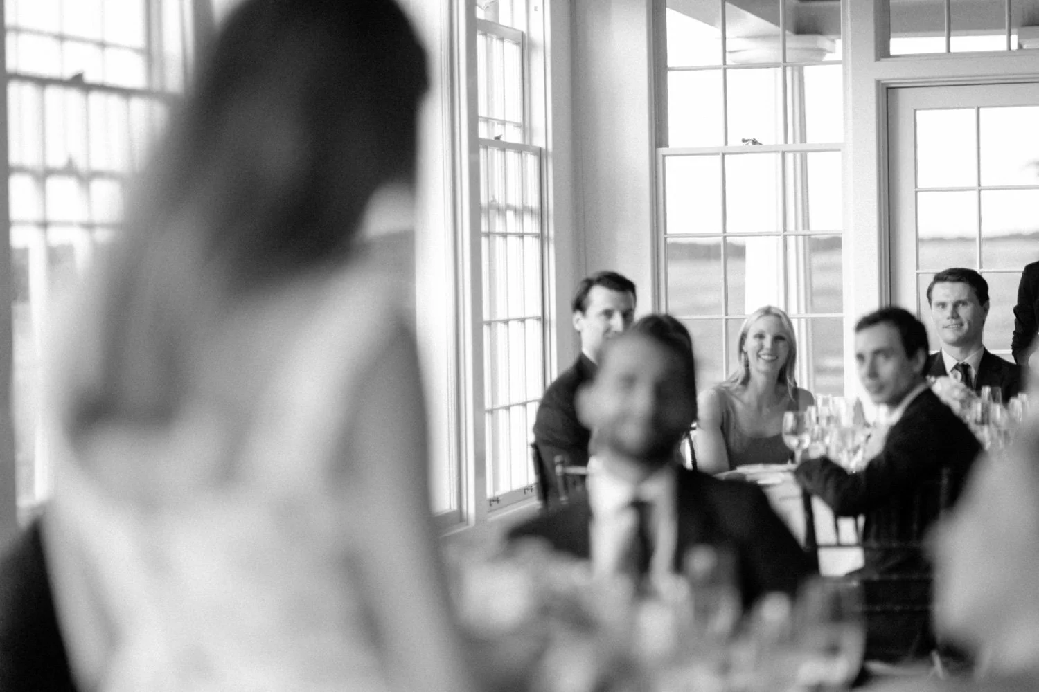 Black-and-white photo of people seated at a table in a bright room with large windows, engaged in a rehearsal dinner at Shinnecock Golf Club.
