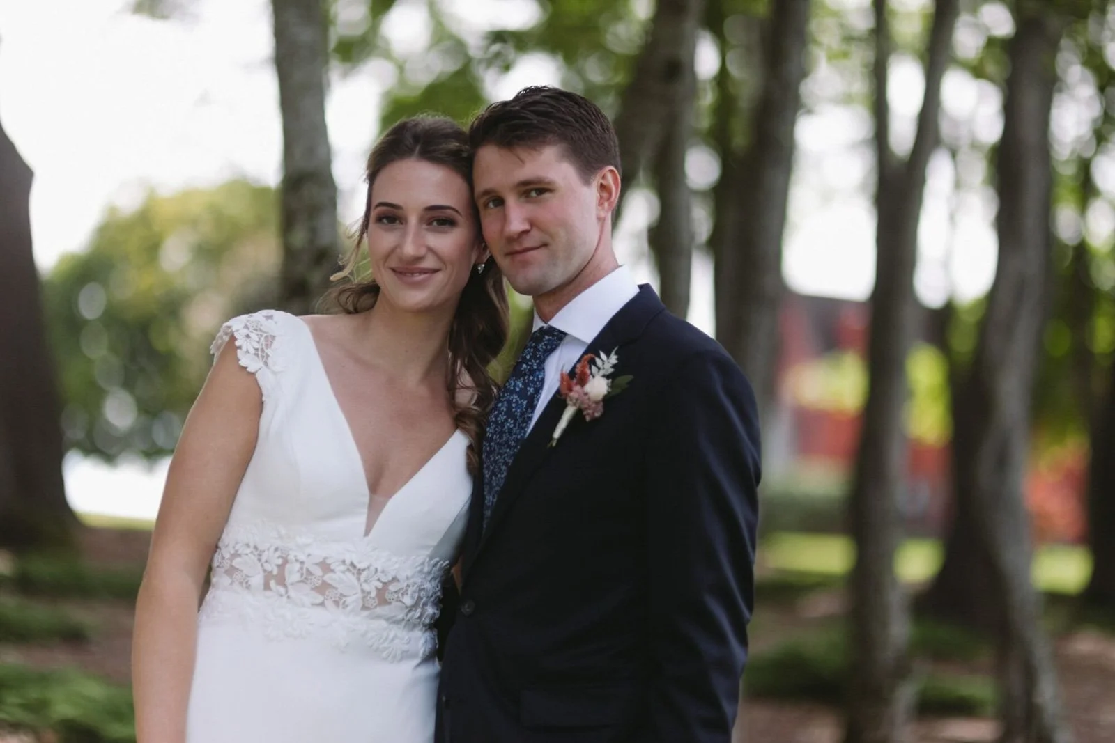 A bride and groom stand close together outdoors, smiling at the camera. The bride wears a white dress with lace details and the groom wears a dark suit with a white shirt and patterned tie, with a boutonniere on his lapel. Forest trees are in the bac