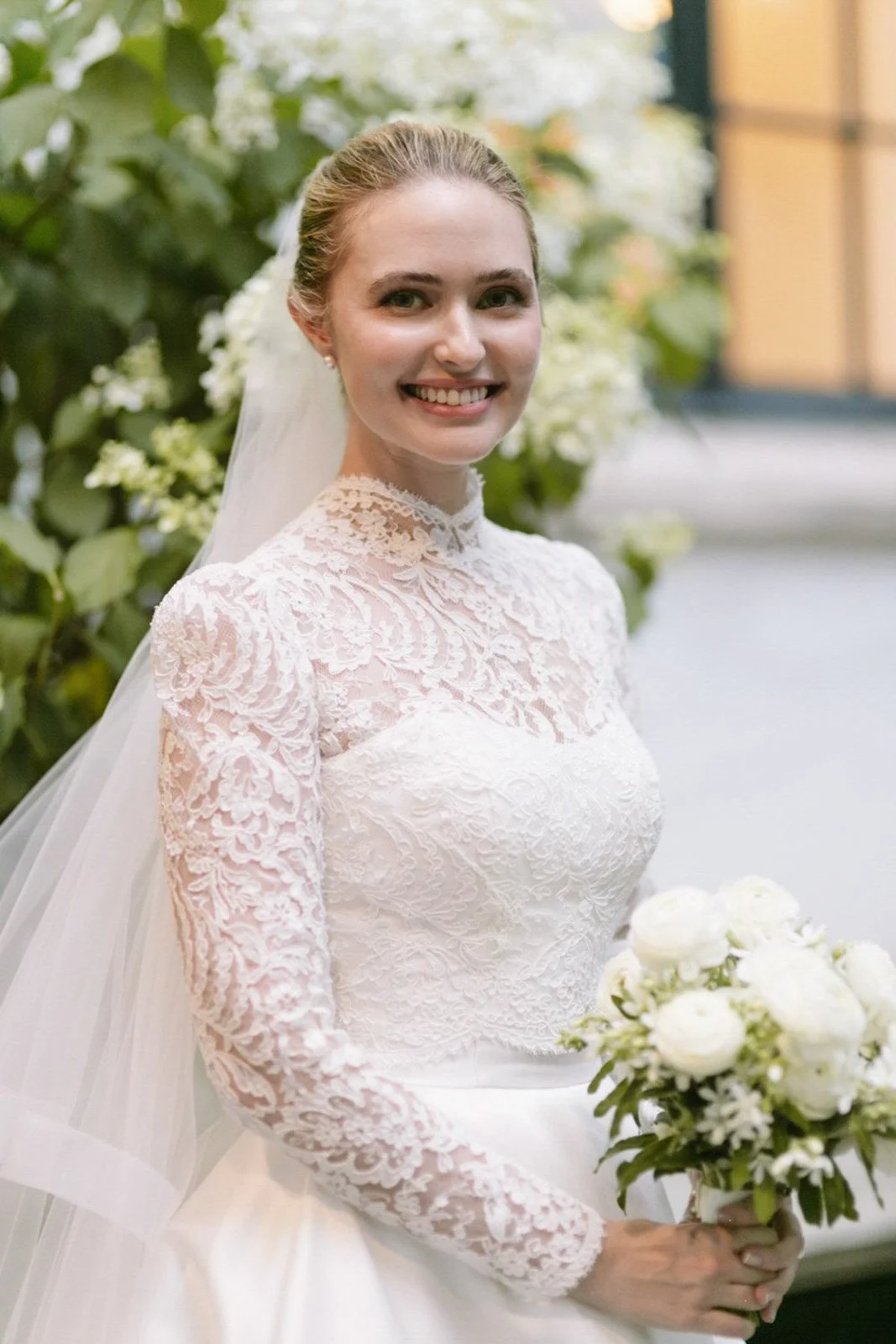 Bride in a lace wedding dress holding a bouquet of white flowers, smiling indoors with greenery background.