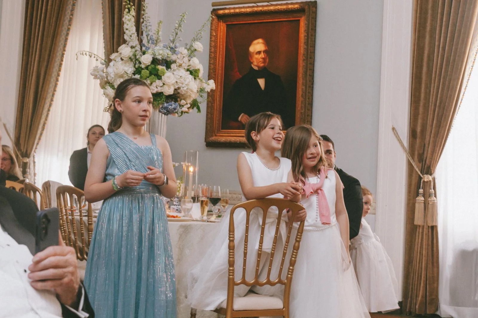 Children standing and sitting at a formal event in a decorated room with a large portrait and floral arrangements.