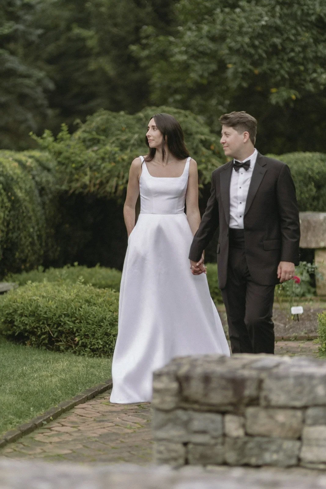 A bride and groom holding hands while walking outdoors in a garden setting, with the bride wearing a white wedding dress and the groom in a black tuxedo.