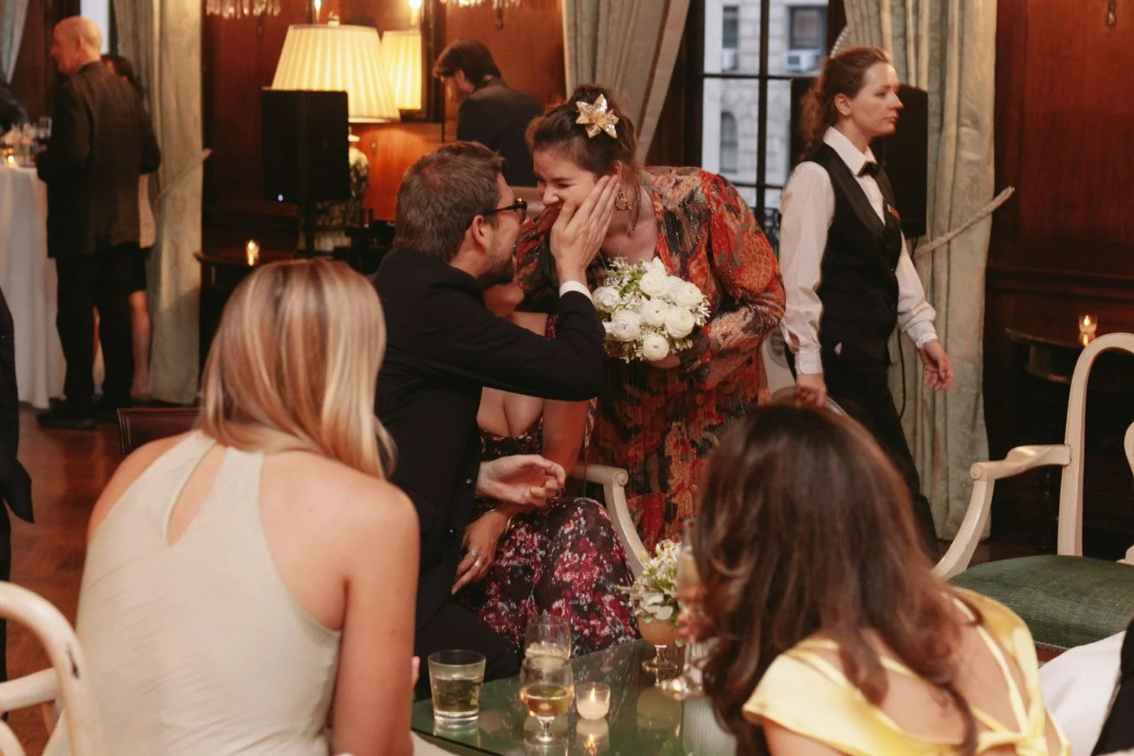 A woman in a colorful dress holding a bouquet of white flowers is surprised and happy as a man in glasses and dark suit leans in toward her, touching her face during a social gathering in a warmly lit room.