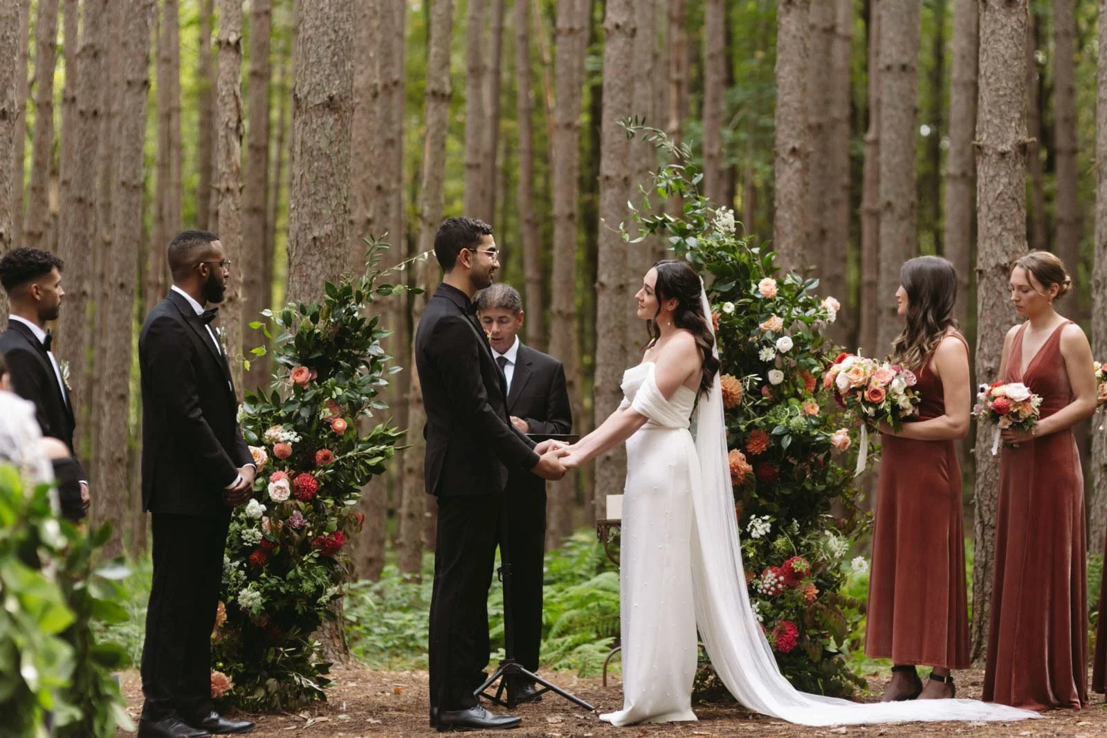 A wedding ceremony taking place in a forest, with the couple holding hands, surrounded by their bridesmaids and groomsmen, floral arrangements, and tall trees in the background.