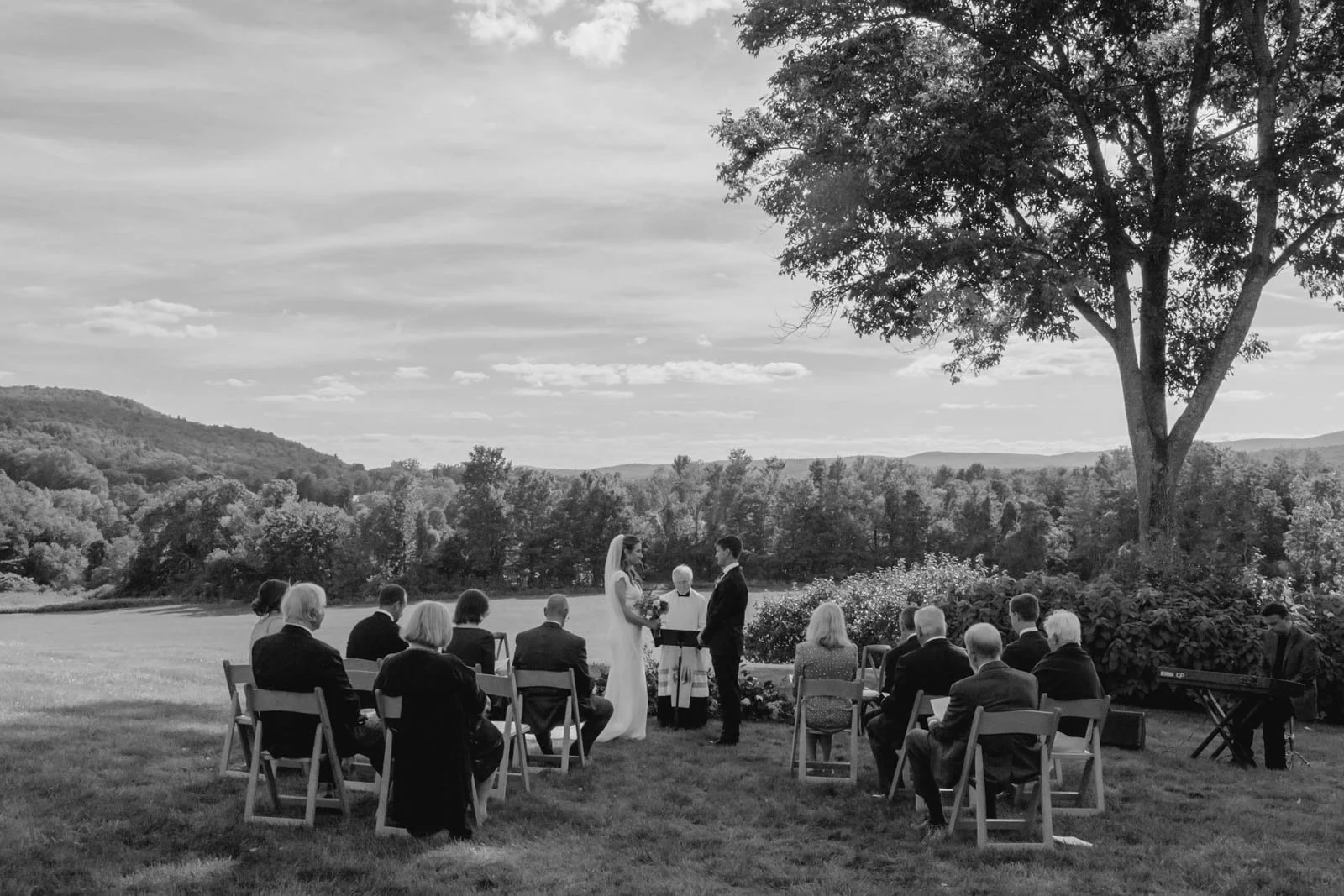 An outdoor wedding ceremony with a bride and groom standing under a tree, surrounded by seated guests on a grassy field, with scenic hills and clouds in the background.