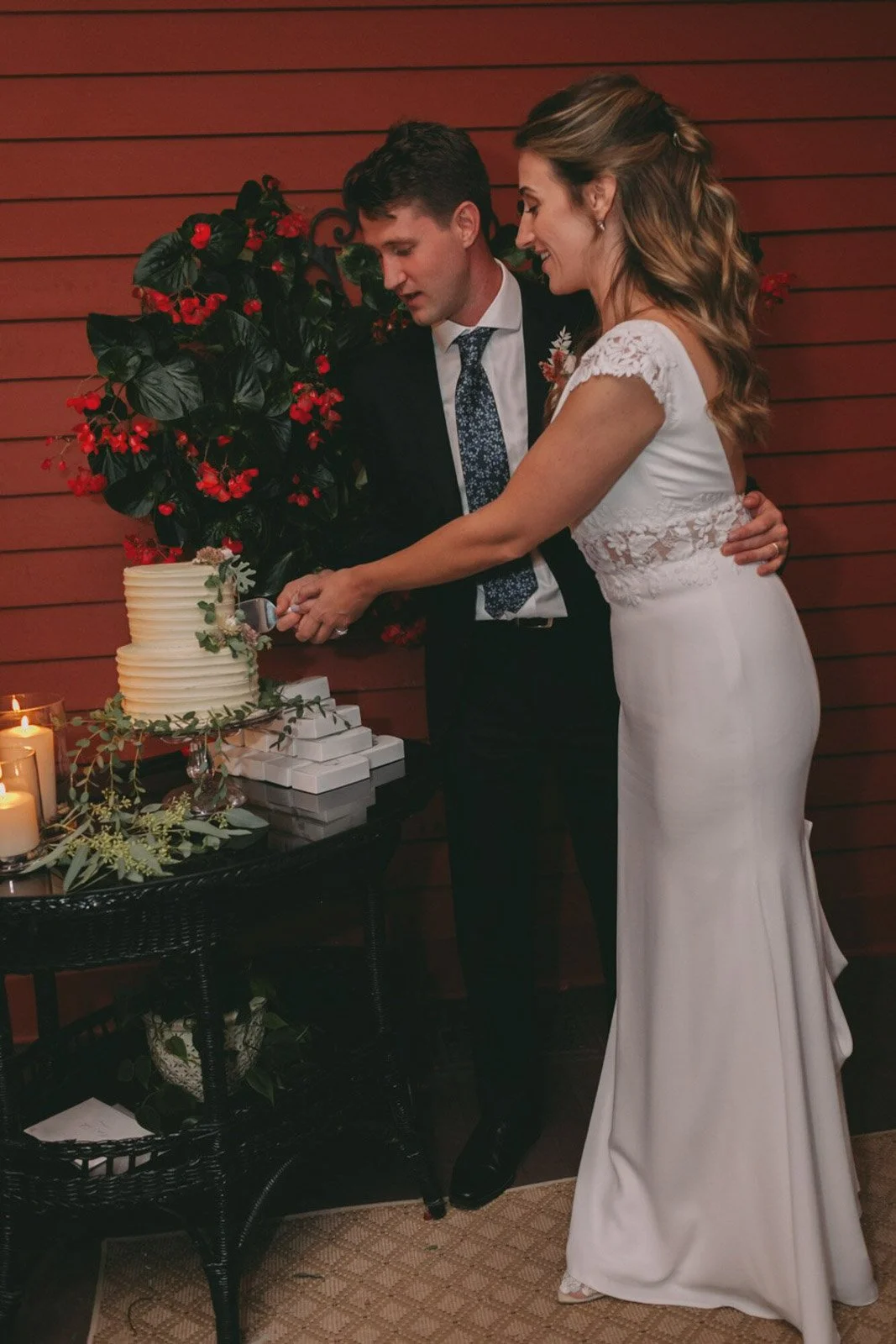 A bride and groom cutting a wedding cake together in front of a red wall with a large plant. The bride is in a white gown, and the groom is in a dark suit with a tie.
