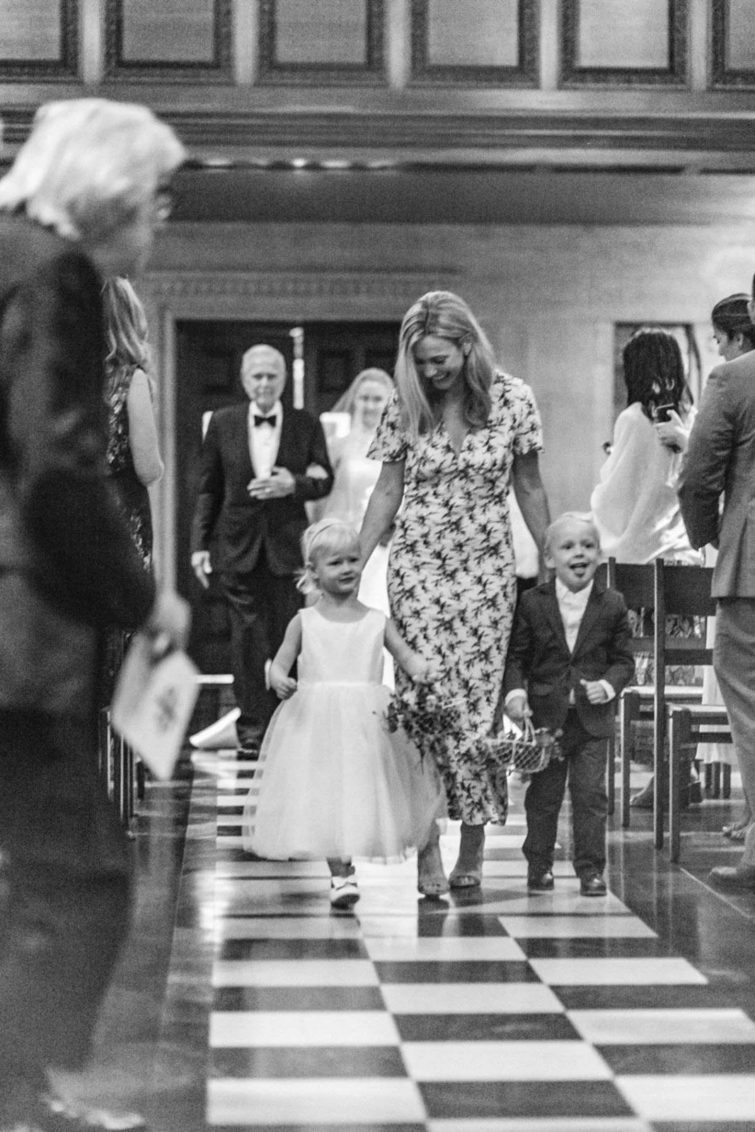 A woman in a floral dress walking down an aisle with two young children, a girl in a white dress and a boy in a suit, during a wedding ceremony in a church.