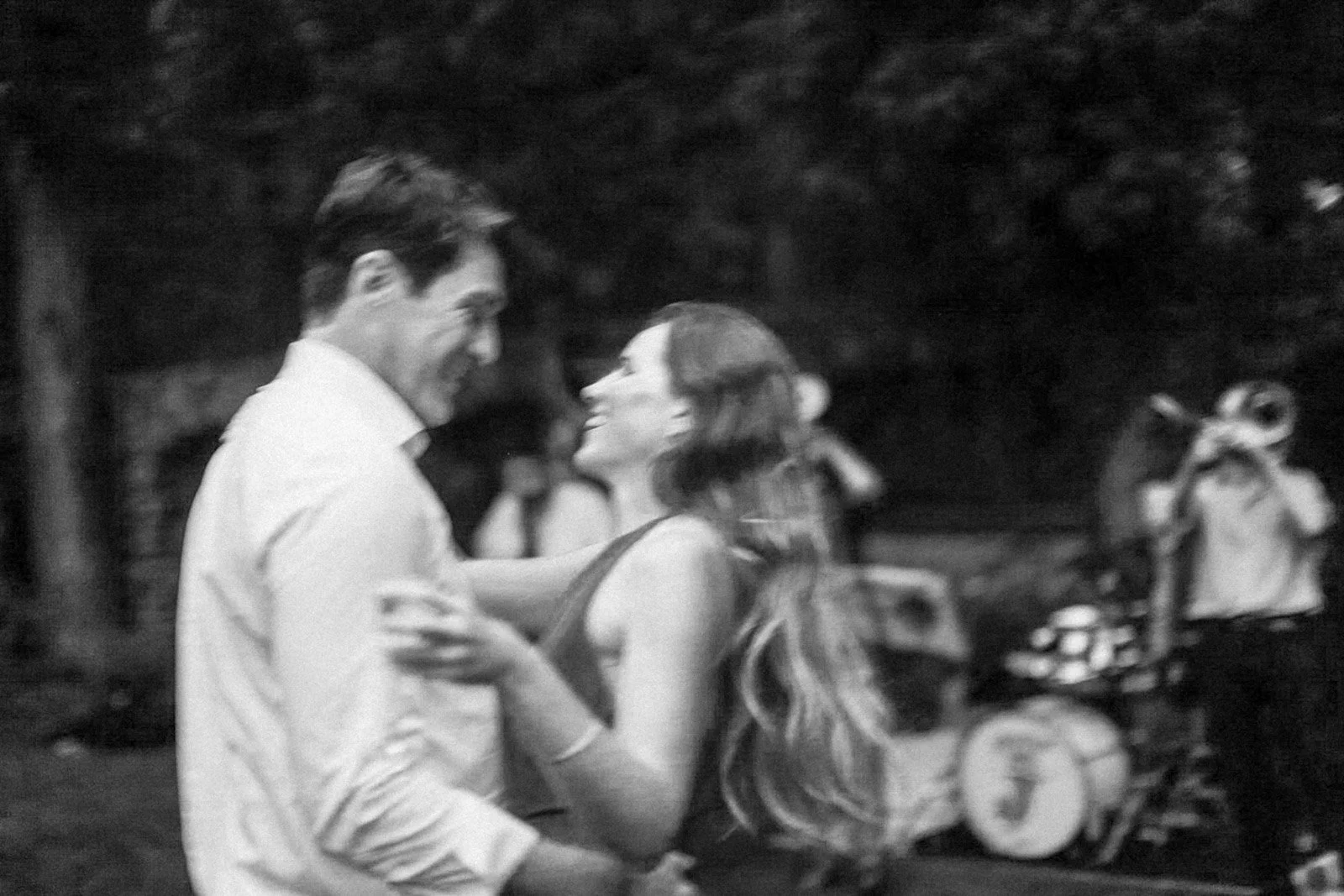 A black and white photo of a couple dancing outdoors at night, smiling at each other with blurred other people in the background.