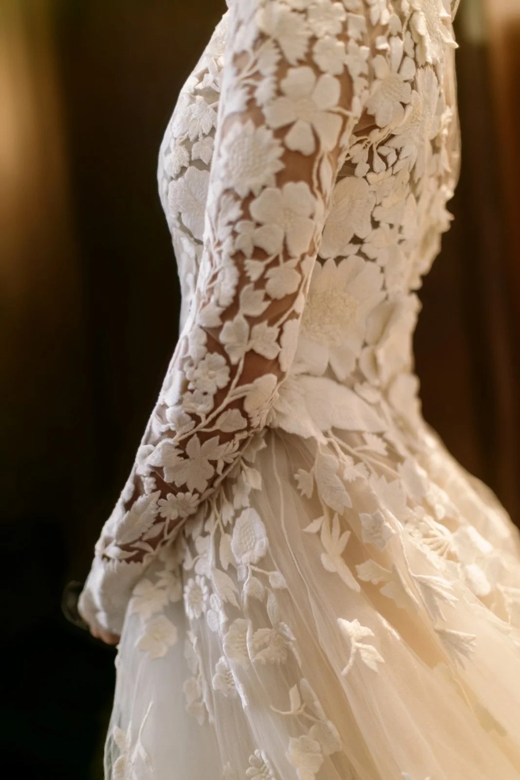 Close-up of a wedding dress with floral lace and intricate embroidery, showing the long sleeve and part of the skirt.