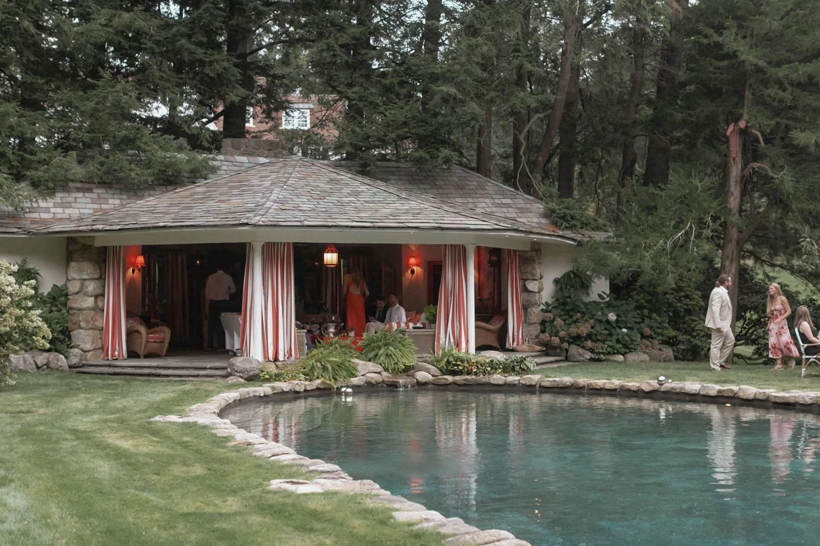 A group of people socializing outdoors near a house with a shaded patio, overlooking a swimming pool, surrounded by tall trees and lush greenery.