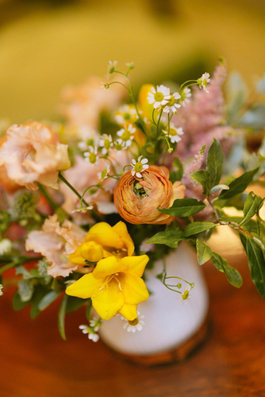 A bouquet of various flowers in a white vase, including yellow, pink, orange, and white blossoms with green leaves, on a wooden surface.