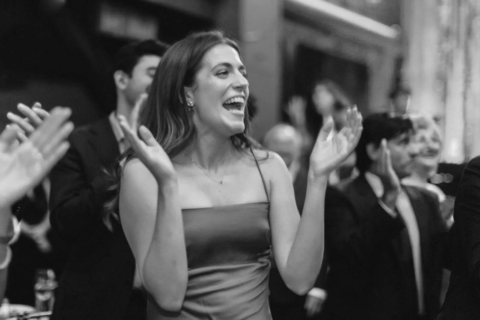 A woman in a strapless dress is smiling and clapping at an indoor event, surrounded by other people.