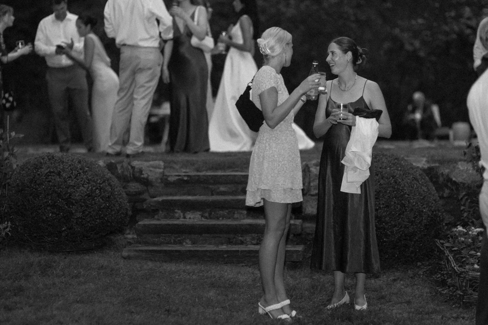 Two women standing and talking at an outdoor gathering, holding drinks, with other people in the background.