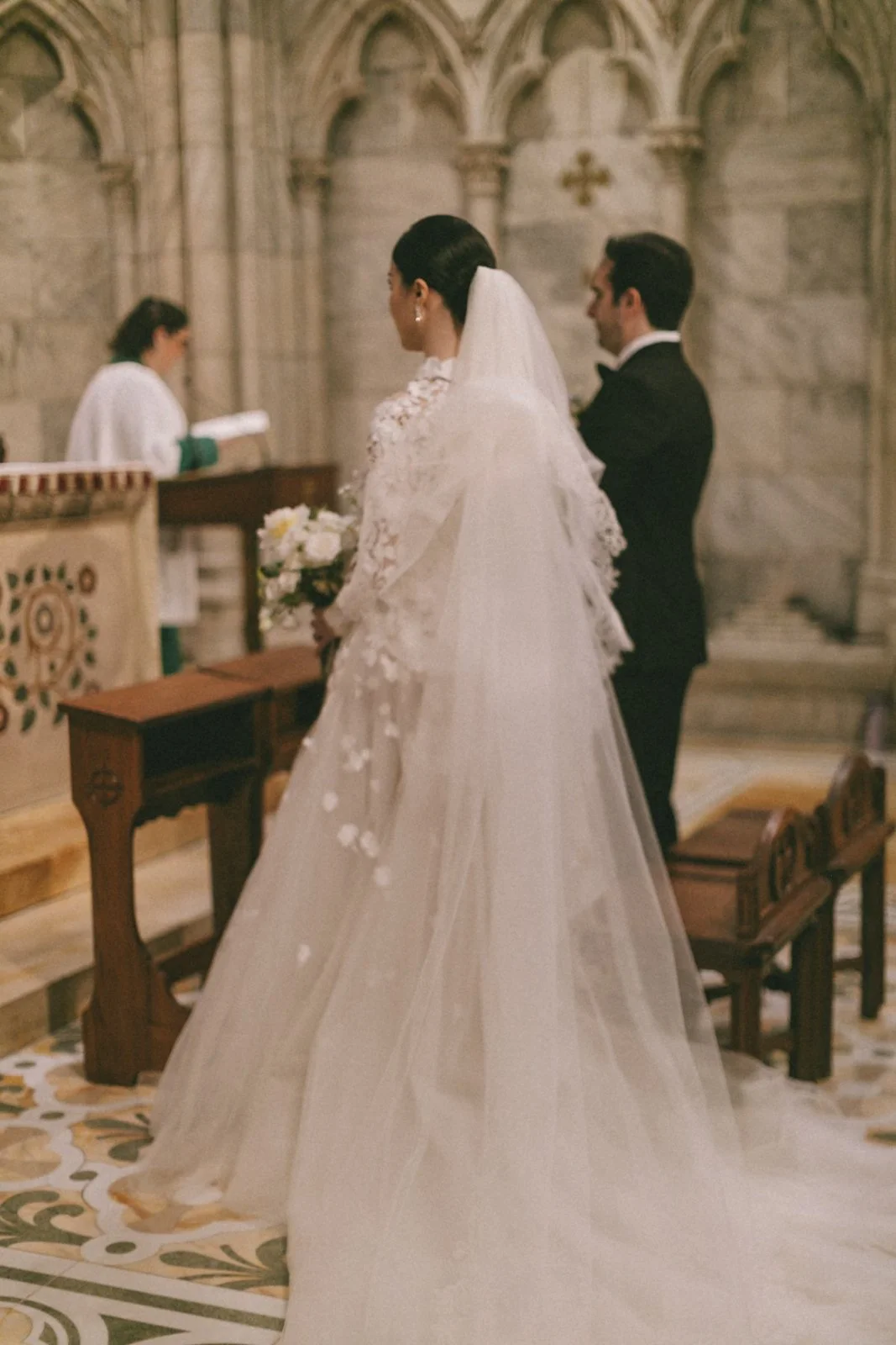 A bride and groom standing at the altar during a wedding ceremony inside a church with traditional stone architecture, a priest officiating, and floral details.
