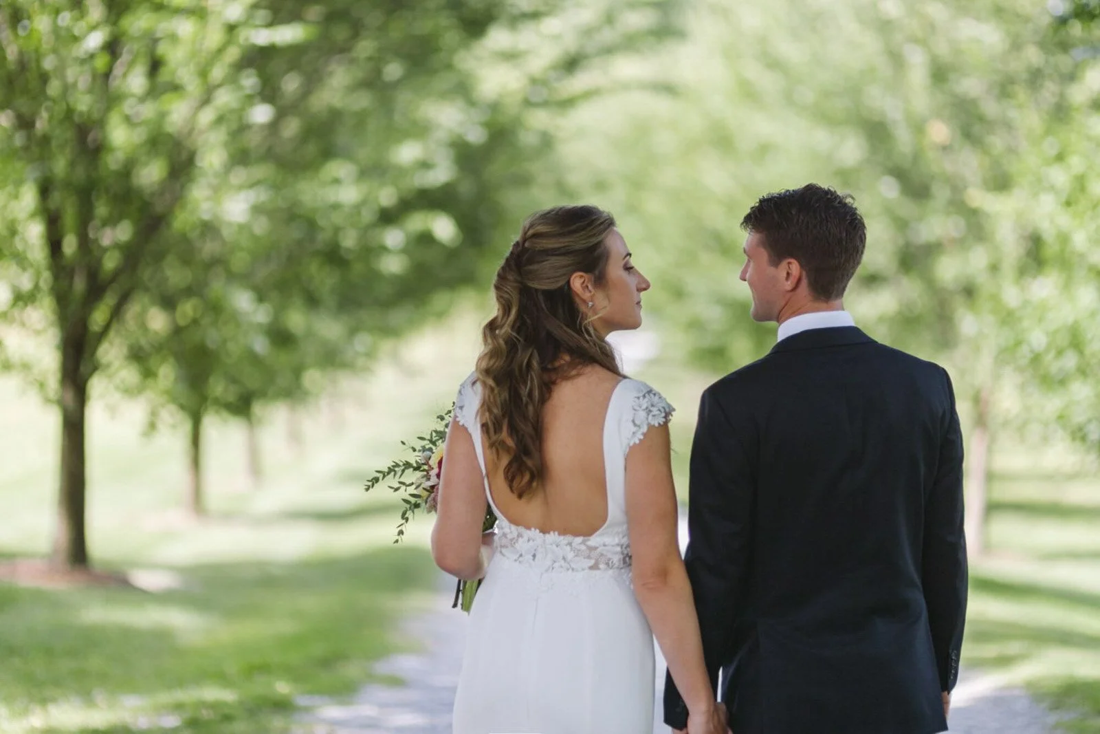 A bride and groom holding hands in a park with green trees in the background, facing each other at Court Hill in The Berkshires.