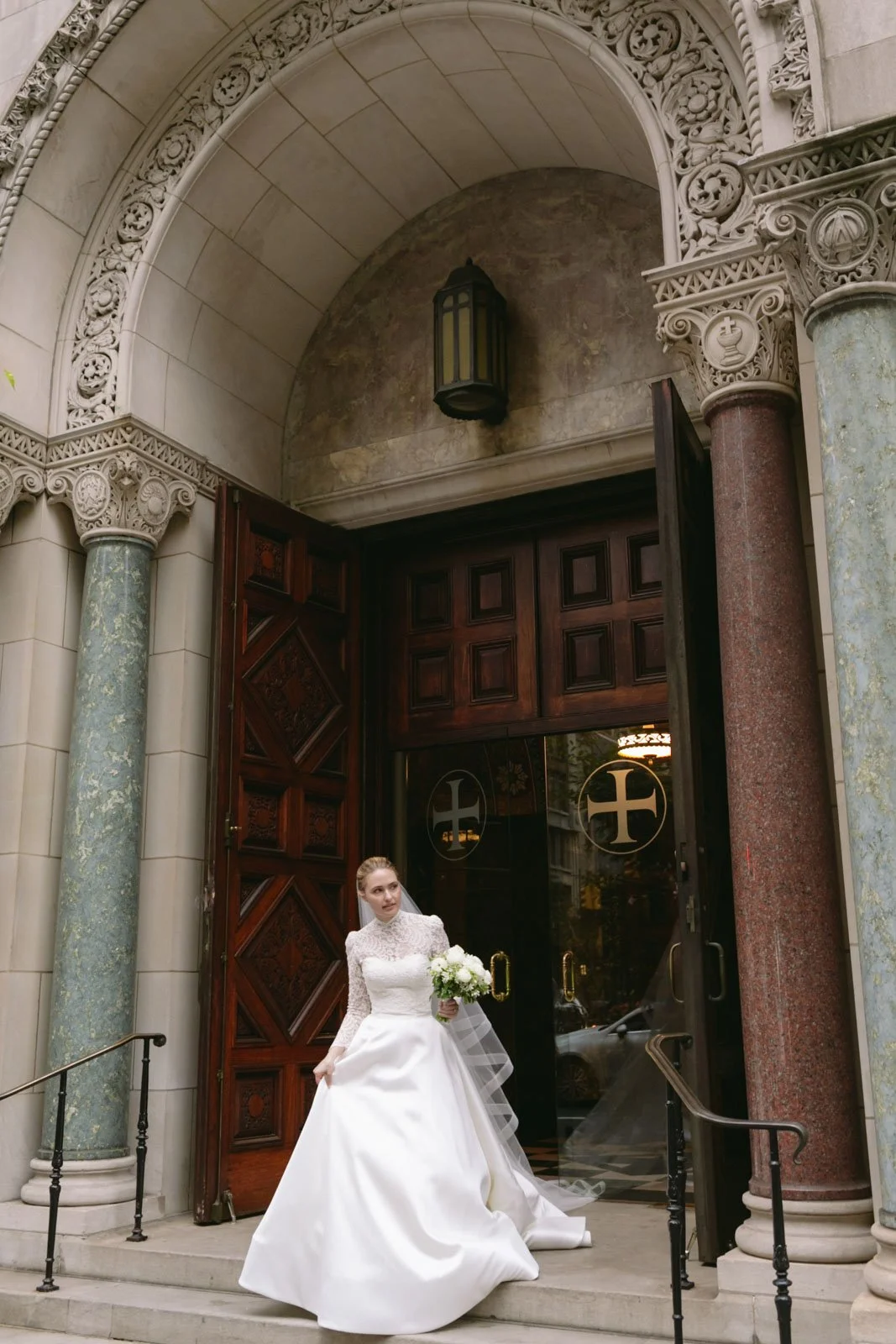 Bride in a white wedding dress holding a bouquet, standing in front of Christ Church NYC with ornate columns and wooden doors.
