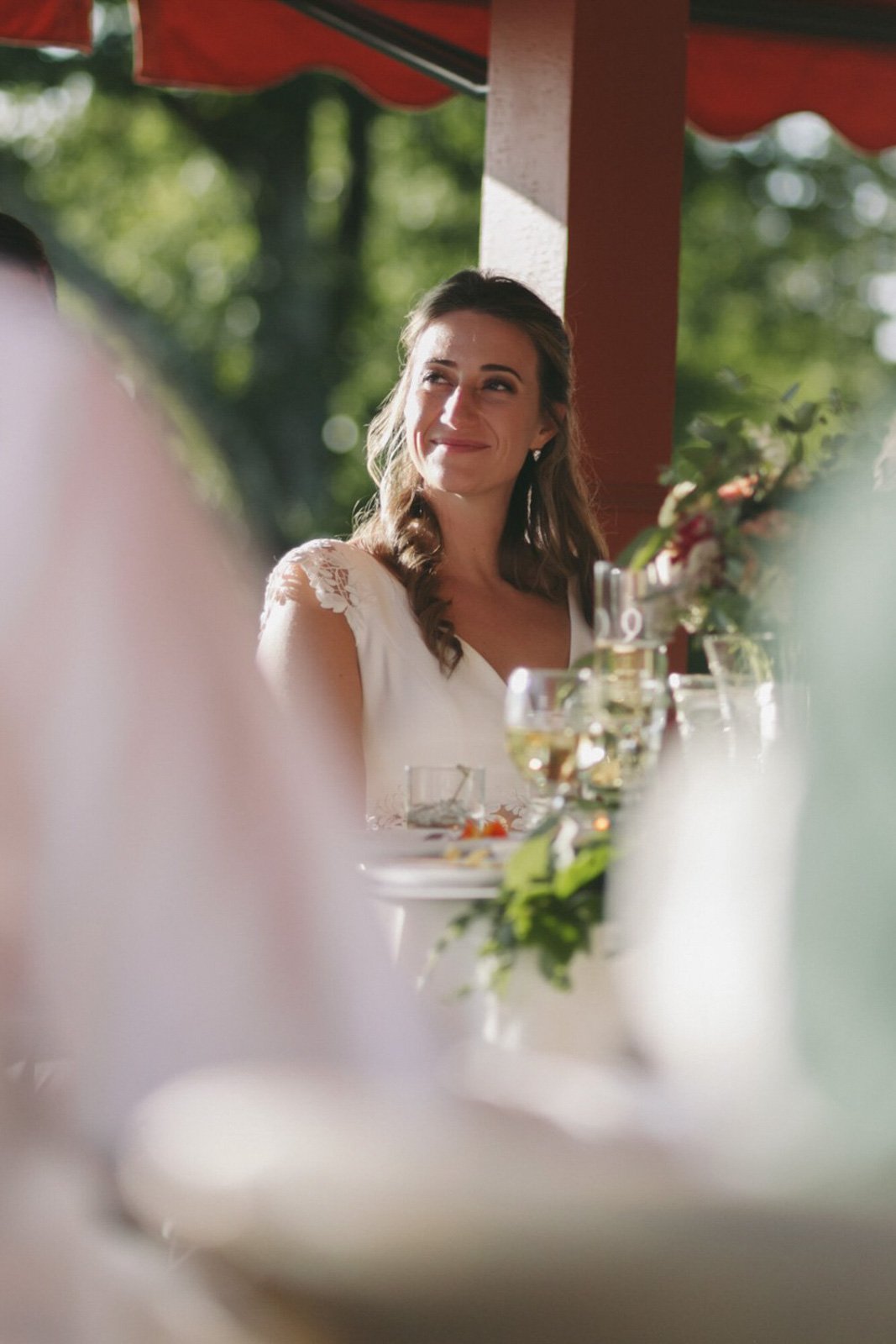 A woman with long wavy brown hair, wearing a white dress with lace shoulder details, smiling and looking to her right, at an outdoor gathering with a blurred background of green trees and tables with glasses and plates.