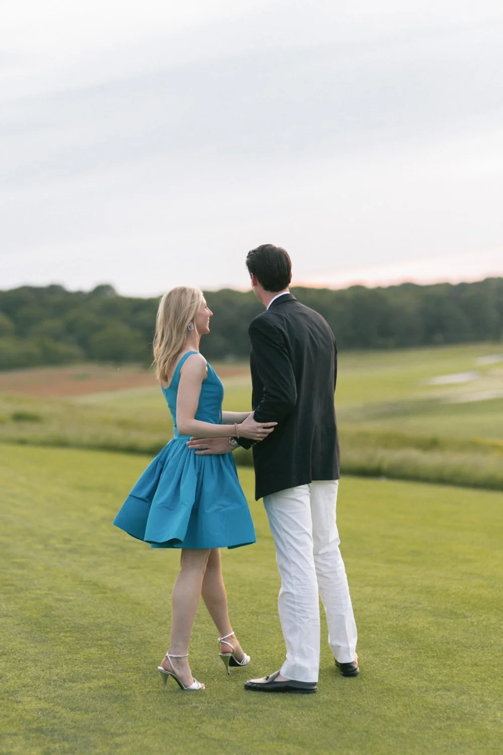 A couple dressed in semi-formal attire standing on a grassy field, holding hands and facing each other, with a scenic background of trees and an overcast sky.