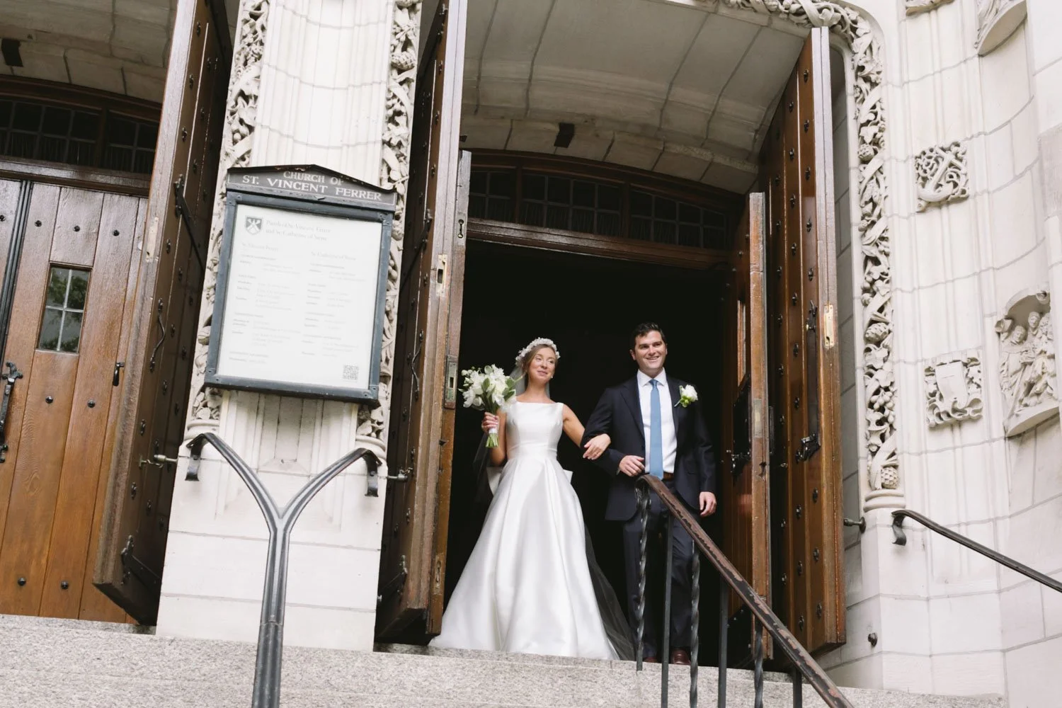 A bride and groom exiting the church of St. Vincent Ferrer on their wedding day. The bride is holding a bouquet of white flowers, wearing a white wedding dress, and the groom is in a dark suit, white shirt, and tie, smiling as they walk down the step