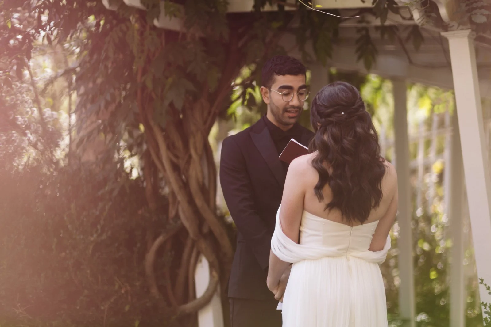 A couple at an outdoor wedding ceremony with a man reading vows to a woman dressed in a strapless white wedding gown, surrounded by greenery and trees at the Roxbury Barn and Estate.
